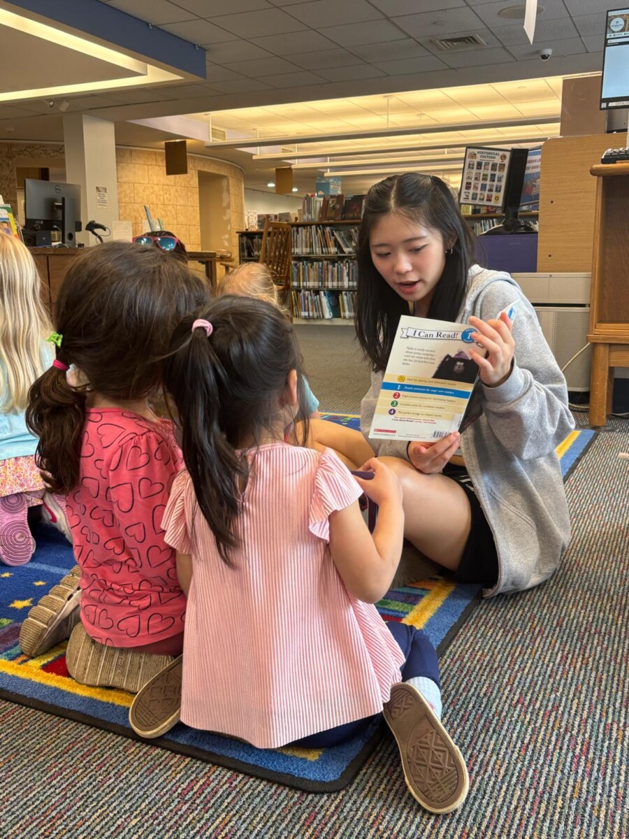 Seated on the floor, a young person reads to a group of children at the library